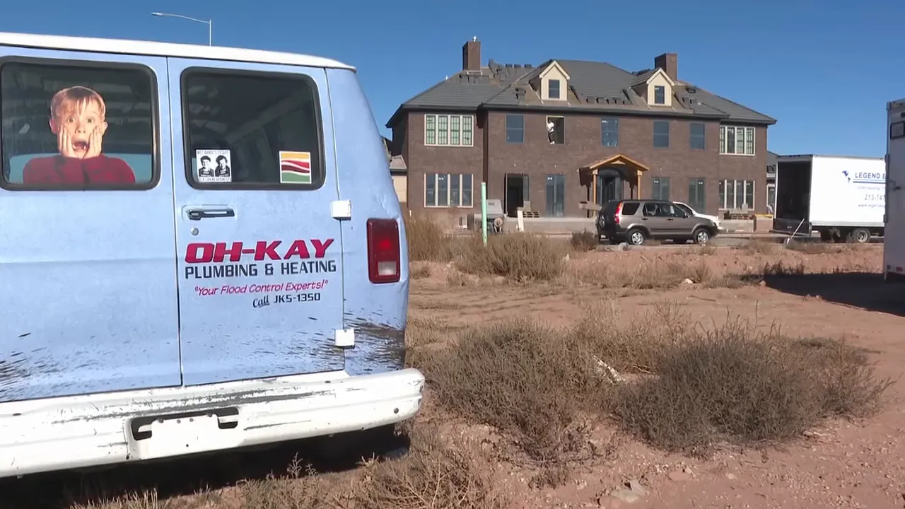 A light-blue plumbing van with a Home Alone–style window decal parked in front of the brick house under construction.